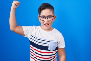 Wall Mural - Young hispanic kid standing over blue background angry and mad raising fist frustrated and furious while shouting with anger. rage and aggressive concept.