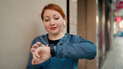 Wall Mural - Young redhead woman wearing sportswear listening to music looking stopwatch at street