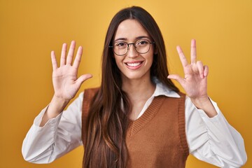 Sticker - Young brunette woman standing over yellow background wearing glasses showing and pointing up with fingers number eight while smiling confident and happy.