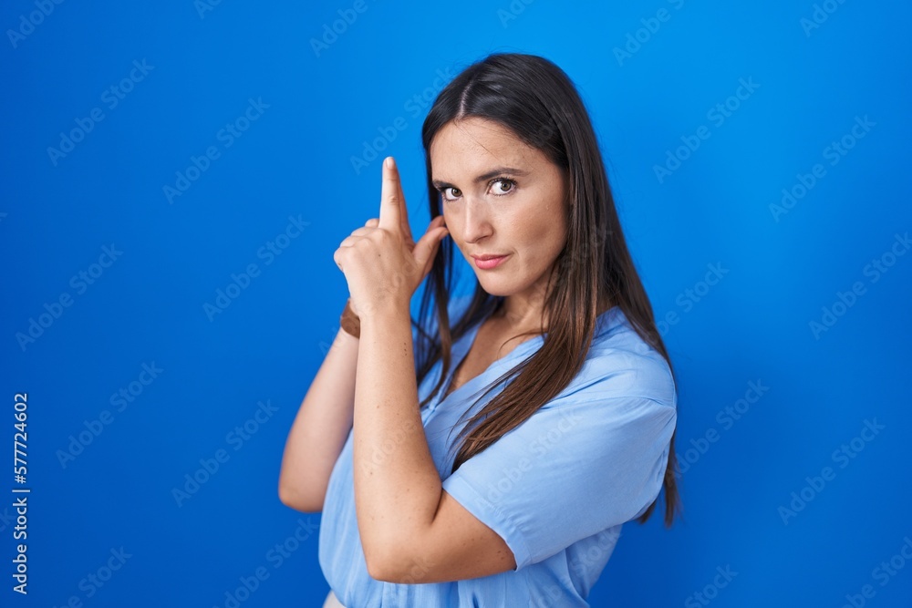 Young brunette woman standing over blue background holding symbolic gun with hand gesture, playing killing shooting weapons, angry face