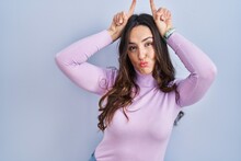 Young Brunette Woman Standing Over Blue Background Doing Funny Gesture With Finger Over Head As Bull Horns