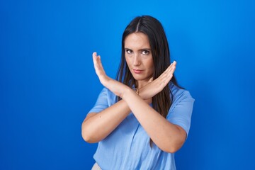 Sticker - Young brunette woman standing over blue background rejection expression crossing arms doing negative sign, angry face