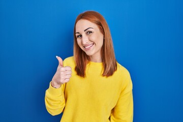 Poster - Young woman standing over blue background doing happy thumbs up gesture with hand. approving expression looking at the camera showing success.