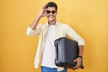 Wall Mural - Young hispanic man holding suitcase going on summer vacation doing ok gesture with hand smiling, eye looking through fingers with happy face.