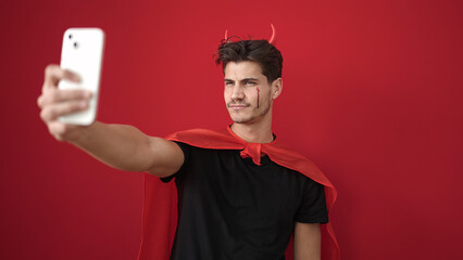 Wall Mural - Young hispanic man wearing devil costume using smartphone over isolated red background