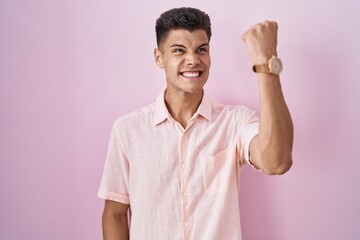 Sticker - Young hispanic man standing over pink background angry and mad raising fist frustrated and furious while shouting with anger. rage and aggressive concept.