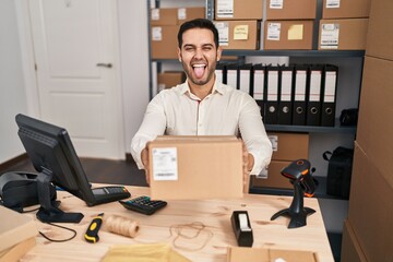 Poster - Young hispanic man with beard working at small business ecommerce holding box sticking tongue out happy with funny expression.