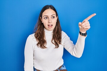 Sticker - Young hispanic woman standing over blue background pointing with finger surprised ahead, open mouth amazed expression, something on the front