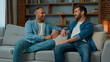 © Yuliia - Two diverse 30s men businessmen talking at home on sofa multiracial friends companions african american caucasian ethnicity fellows brothers discussing plans friendly talk male dialogue conversation