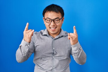 Sticker - Young chinese man standing over blue background shouting with crazy expression doing rock symbol with hands up. music star. heavy concept.