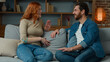 © Yuliia - Woman talking with man at home joyful Caucasian couple sit on couch redhead girl telling funny story positive diverse partners laughing with joke having fun laugh discussing news friendly talk on date