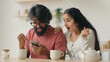 © Yuliia - Multiracial couple indian man and woman sit in kitchen using mobile phone application enjoying web surfing read news win online bonus on smartphone happy ethnic spouses rejoice victory give high five