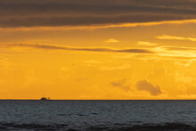 A Boat Floats On The Sea In The Distance At A Bright Yellow Sunset