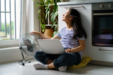 Carefree positive Asian female copywriter working sits in kitchen on floor with laptop and pointing fan at herself. Relaxed Chinese girl enjoys distance work in comfortable. Summertime. 