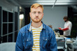 Young red-haired programmer or businessman in modern office looking seriously at camera. Freelancer with arms crossed poses concentrated for photo in meeting room. Colleagues work in the background.