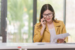 © NanSan - Female attractive asian banker accountant wearing yellow formal suit calculate the cash bill of audit and balance sheet working with quarterly reports of company. call a mobile phone