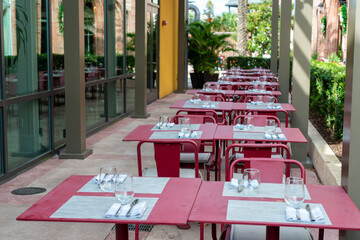 The exterior patio area of a small restaurant entrance with empty retro chairs, benches, and wooden tables. The building has glass windows and a door. The tables are bright red and set with utensils. 