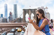 © Maridav - Phone. New York City woman using phone app walking on Brooklyn Bridge by Manhattan city skyline. Young female professional multicultural lady wearing sunglasses, New York City, USA.