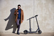 © baranq - Handsome man standing in urban setting with electric scooter using smartphone