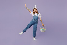 Full Body Smiling Fun Young Woman Wearing Casual Clothes Bunny Rabbit Ears Holding Wicker Basket Colorful Eggs Raising Hand Up Isolated On Plain Pastel Purple Background Studio. Happy Easter Concept.