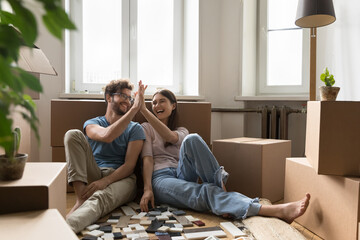 Cheerful excited young couple planning renovation after moving into new apartment, choosing interior material samples of tile, laughing, giving high five, clapping hands at cardboard boxes