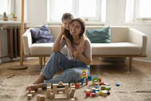 Joyful Sweet Child Embracing Happy Pretty Young Mom Sitting On Floor At Toy Castle. Positive Mother And Cheerful Kid Looking At Camera, Posing, Playing Game Wit Building Blocks At Home