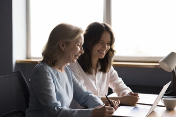 Happy cheerful different aged office colleagues women talking at shared laptop, looking at screen, display, talking on video call, watching online project presentation, laughing