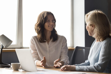 Wall Mural - Two cheerful different aged business colleagues chatting at work table, discussing work success, smiling, laughing. Positive young employee consulting mature mentor woman