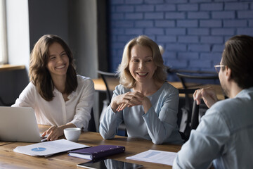 Wall Mural - Happy different aged team of office coworkers meeting at table, talking, laughing, discussing project. Senior professional, expert giving consultation to cheerful young employees, speaking, joking