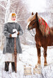 Russian beauty girl in traditional clothes stands with a yoke and buckets. Behind red big horse in forest winter