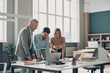 © gstockstudio - Three confident business people looking at laptop while standing near the office desk together