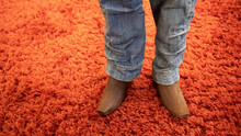 Standing And Walking Legs Of A Wooden Puppet Or Marionette Dressed In Cloth Of Jeans And Boots Of Wood, On A Red Carpet In The Background
