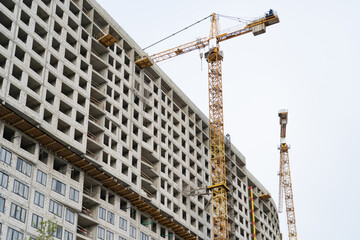 Yellow construction tower cranes and unfinished residential building against grey sky. Housing construction, apartment block with scaffolding