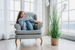 © Iona - Attractive blonde American young woman sitting in cozy chair hugs bended legs looks at camera smiles loves her apartments with large  windows. Relaxed fashion model in casual enjoys weekend home.