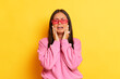 Indoor shot of hesitant brunette woman shrug shoulders put hands on the face cry and doubt while make decision stands puzzled wears modern pink sweater and pink glasses isolated over yellow background