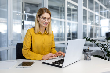 happy and successful business woman working inside office with laptop, female worker in yellow shirt
