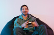 © arthurhidden - Positive young man in casual clothes sitting on pouf using joystick while playing video game in room illuminated by neon lights looking at camera