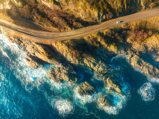 Poster - Aerial view of road, rocky sea coast with waves and stones at sunset in Lofoten Islands, Norway. Landscape with beautiful road, transparent blue water, rocks. Top view from drone of highway in summer