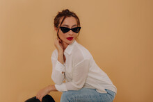 Lovely Cute Charming Girl With Dark Hair And Red Lips Wearing White Shirt, Jeans And Dark Glasses Posing To Camera During Photoshoot Over Isolated Background