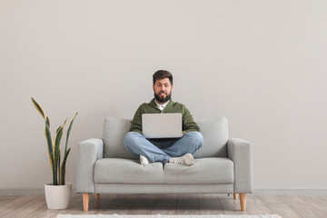 Handsome bearded man using laptop on grey sofa near light wall