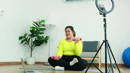 Wall Mural - Asian fat woman streamer sits exercising lifting dumbbells study from trainer on tablet internet online with live streaming video recording on smartphone using ring light brighten up during broadcast.