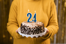 Woman holding birthday cake with burning candles number 24 against decorated background, close up. Celebrates birthday holiday at home.