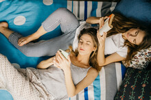 Two Women Twins Are Lying On Bed, Reading A Book And Typing Smartphone