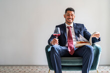 Happy Businessman Sitting With Book And Glass Of Red Wine On Armchair In Front Of Wall