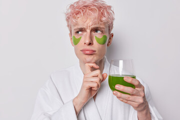 Wall Mural - Photo of displeased thoughtful millennial guy keeps hand under chin looks aside unhappily has sulking expression wears green pads under eyes undergoes morning cosmetic procedures drinks fresh smootie