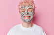 Portrait of cheerful adult man with broad toothy smile keeps eyes closed enjoys skin care beauty procedures applies facial clay mask dressed in casual white t shirt isolated over pink background.
