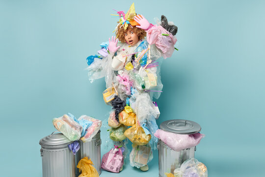 Scared shocked curly haired woman keeps palms raised tries to protect herself from danger wears plastic garbage costume stares impressed at camera surrounded by litter bins busy picking up wastes