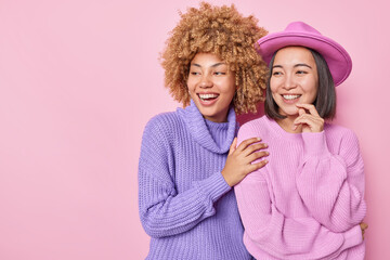 Wall Mural - Horizontal shot of cheerful mixed race women wears knitted jumpers and hat stand closely to each other smile gladfully isolated over pink background blank space for your advertising content.