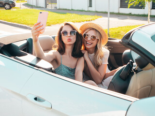 Two young beautiful and smiling hipster female in convertible car. Sexy carefree women driving. Positive models riding and having fun outdoors. Enjoying summer days. Taking selfie photos