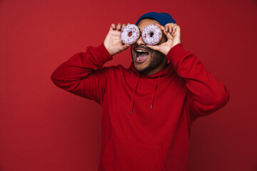 Positive screaming indian man covering his eyes with donuts isolated over red background